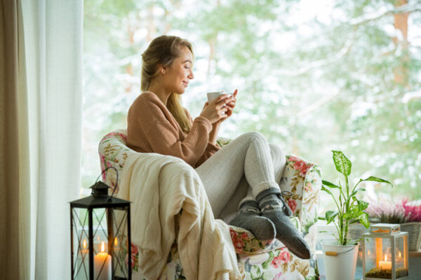 Person sitting on a bed near a window with soft morning light and a warm drink